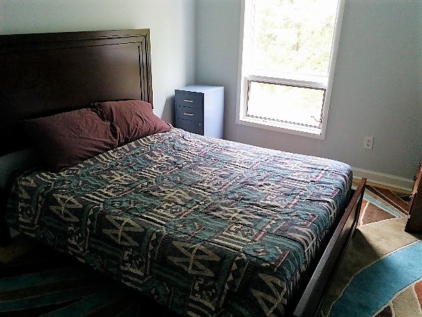 large bed with green and blue multi colored bedspread with wooden headboard in small room with window and filing cabinet